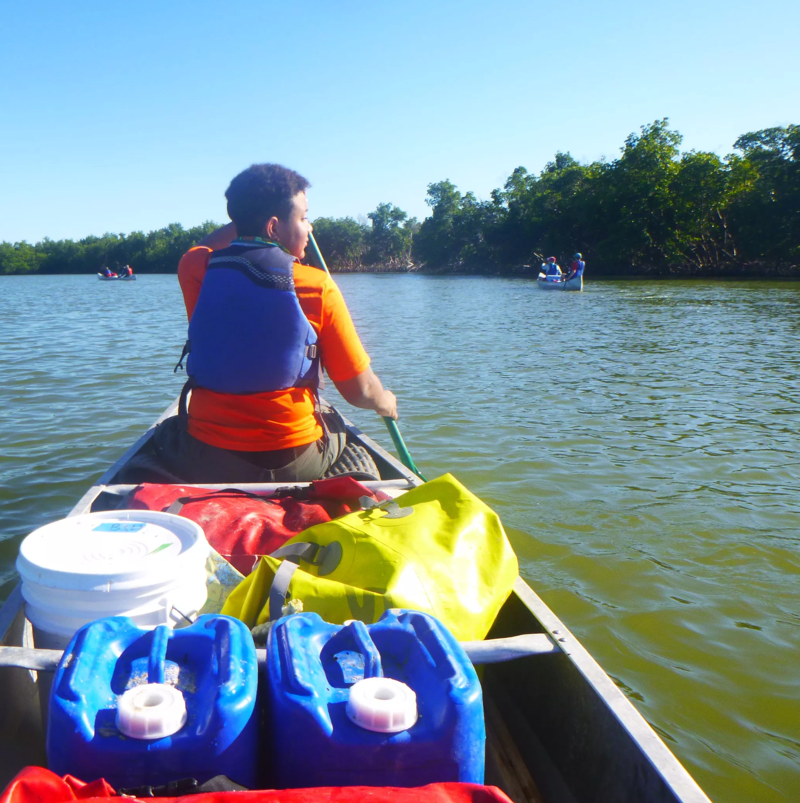 A person wearing an orange shirt and a blue life vest is paddling a canoe on a river. There are blue and white containers in the canoe. Other people in canoes can be seen in the distance. The river is surrounded by green trees and the sky is blue.